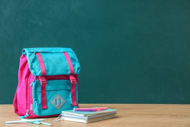 Yeşil karatahtanın yanındaki kahverengi ahşap masada kırtasiye malzemesi olan renkli okul çantası.