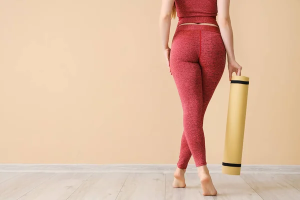 Sporty young woman in leggings and with yoga mat near beige wall, back view
