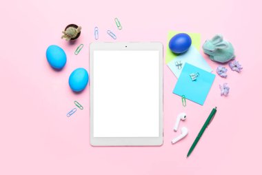 Tablet computer with office stationery, Easter eggs, rabbit and flowers on pink background