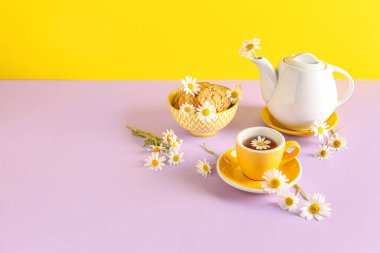 Sarı duvarın yanındaki pembe masada doğal papatya çayı, kurabiye ve çiçeklerle çaydanlık.
