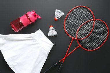 Badminton raketleri, mekik penisleri, bir şişe su ve karanlık arka planda spor kıyafetleri.