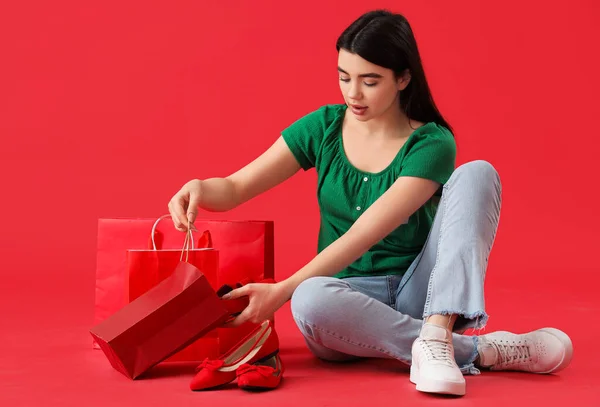 Νεαρή Γυναίκα Κομψά Παπούτσια Και Τσάντες Ψώνια Κάθεται Κόκκινο Φόντο — Φωτογραφία Αρχείου