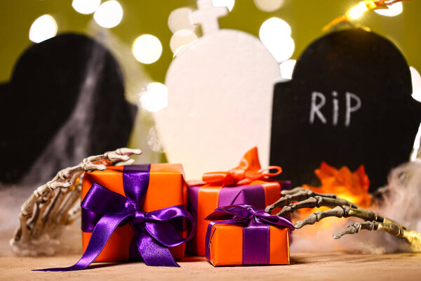 Composition with beautiful gift boxes, skeleton hands and graves for Halloween on table against blurred lights, closeup