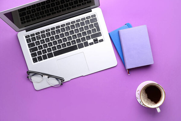 Modern laptop, notebooks, eyeglasses and cup of coffee on purple background