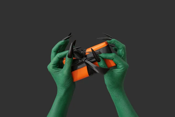 Green hands of witch with gift box on dark background. Halloween celebration