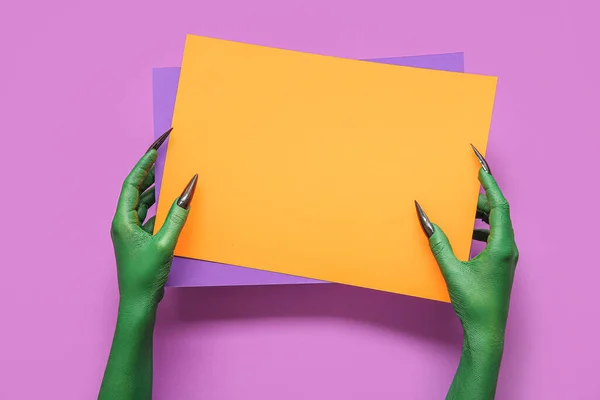 Green hands of witch with blank paper sheets on purple background. Halloween celebration