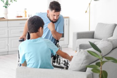 Tekerlekli sandalyede düşünceli bir baba ve küçük oğlu evde satranç oynuyor.