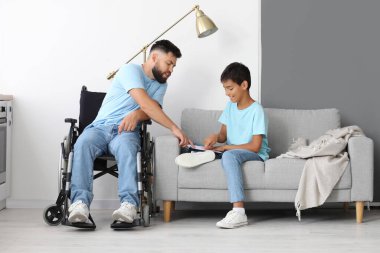 Küçük çocuk ve babası tekerlekli sandalyede evde kitap okuyorlar.