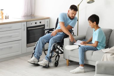 Küçük çocuk ve babası tekerlekli sandalyede evde kitap okuyorlar.