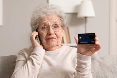 Kredi kartıyla şok olmuş yaşlı bir kadın evdeki koltukta cep telefonuyla konuşuyor.