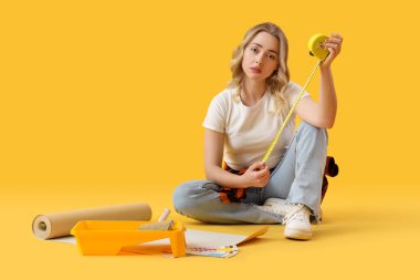 Sarı arka planda mezurası ve duvar kağıdı rulosu olan üzgün genç bir kadın.