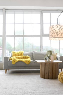 Işıl ışıl oturma odasının içinde rahat gri kanepe, sehpa ve büyük pencerenin yanında parlayan lamba.