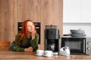 Modern mutfakta kahve makinesi, mikrodalga fırın ve mikseri olan genç kızıl saçlı kadın.