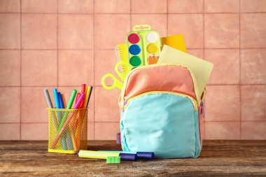 Renkli okul çantası, içinde not defterleri, suluboya resimler ve bej kiremit duvarın yanındaki ahşap masa üzerinde bir fincan işaretleyici.