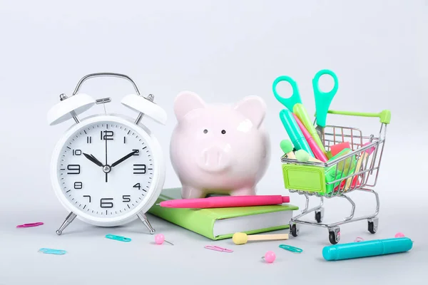 Alarm clock, shopping cart full of different stationery and piggy bank on white background