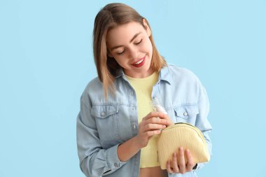 Mavi arka planda makyaj malzemelerini makyaj çantasına koyan genç bir kadın.