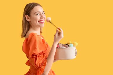 Kozmetik çantası ve sarı arka planda makyaj fırçası olan genç bir kadın.