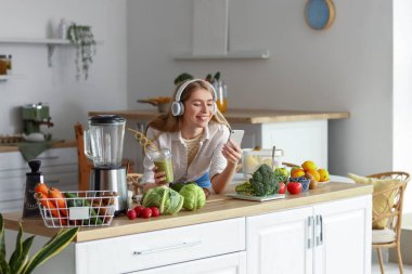 Kulaklık takan, evinde bir bardak sağlıklı meyve suyu ve cep telefonu olan genç bir kadın.
