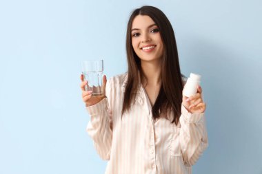 Üzerinde ilaç şişesi ve mavi arka planda bir bardak su olan pijamalı genç bir kadın.