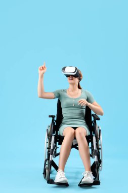 Tekerlekli sandalyedeki genç kadın mavi arka planda VR gözlüğü kullanıyor.