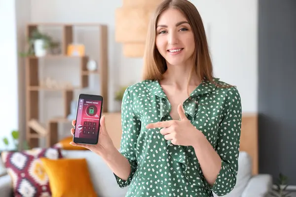 Young woman pointing at mobile phone with smart home security system control panel