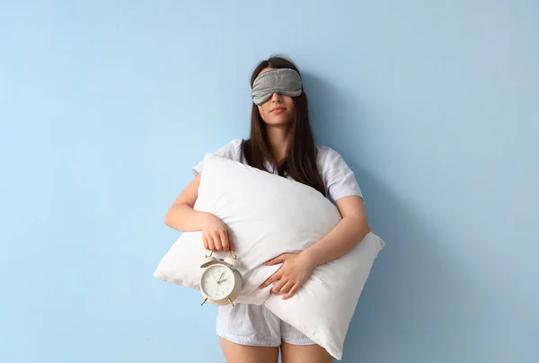 Young woman in sleeping mask with alarm clock and pillow on blue background