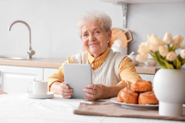 Mutfaktaki masada tablet bilgisayar kullanan yaşlı bir kadın.
