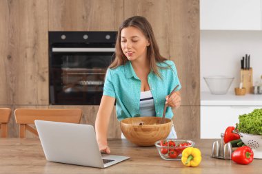 Dizüstü bilgisayarlı genç bir kadın mutfakta sebze salatası hazırlıyor.