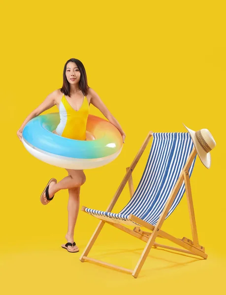 Beautiful Asian woman with swim ring and deck chair on yellow background