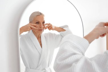 Olgun kadın banyoda aynanın yanında yüz geliştirme egzersizi yapıyor.