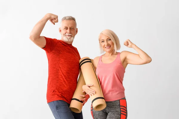 Sporty mature couple with fitness mats showing muscles on light background