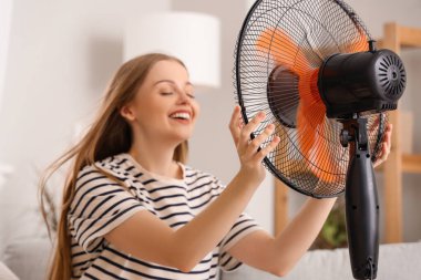 Evde elektrikli vantilatörü olan genç bir kadın.