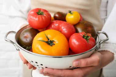 Beyaz tuğlalı arka planda farklı taze domatesli süzgeç tutan kadın, yakın plan.