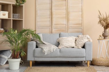 Aydınlık oturma odasının içinde şık gri kanepe, raf ünitesi, katlanır ekran ve çiçekli