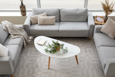 Aydınlık oturma odasının içinde şık gri koltuklar, masa ve çiçek var.