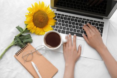 Modern dizüstü bilgisayarı olan kadın eller, güzel ayçiçeği, gözlük ve hafif kumaş arka planda bir fincan kahve.