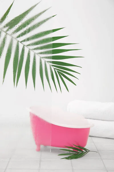 Small bathtub with foam, clean towels and palm leaves on light tile table
