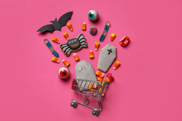 Shopping cart with tasty candy corns and cookies for Halloween on pink background
