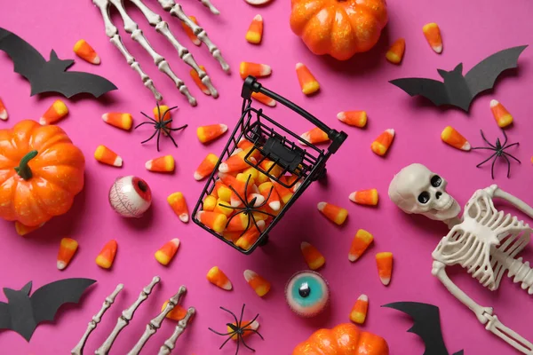 Shopping cart with tasty candy corns for Halloween, skeleton and pumpkins on pink background