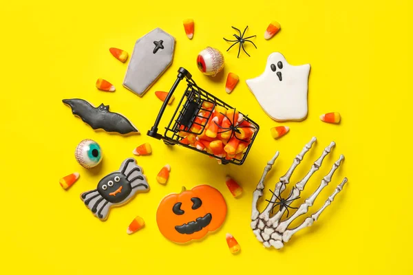 Shopping cart with tasty candy corns, skeleton hand and cookies for Halloween on yellow background