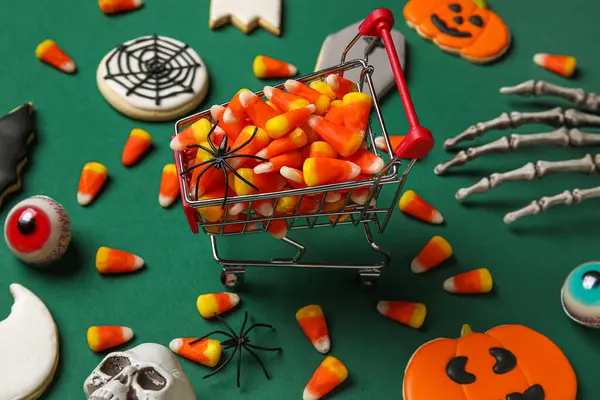 Shopping cart with tasty candy corns, cookies for Halloween and skeleton hand on green background