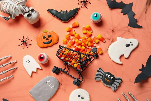 Shopping cart with tasty candy corns, cookies for Halloween and skeleton on red background