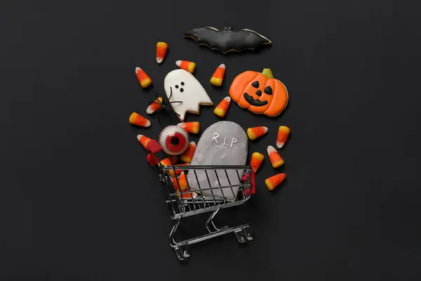 Shopping cart with tasty candy corns, skeleton hand and cookies for Halloween on black background