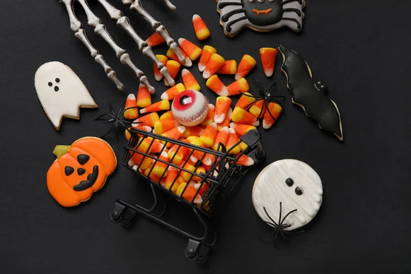 Shopping cart with tasty candy corns, skeleton hand and cookies for Halloween on black background