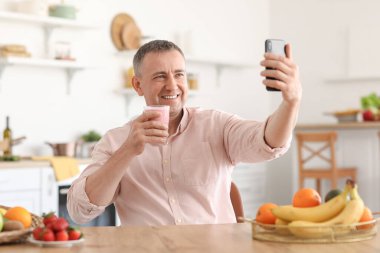 Elinde bir bardak taze meyve suyu olan olgun bir adam mutfakta selfie çekiyor.