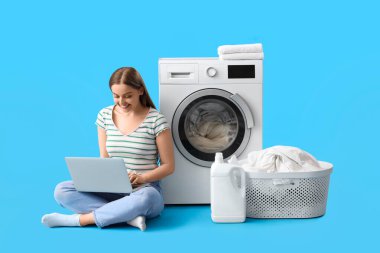 Modern dizüstü bilgisayarı olan güzel bir kadın mavi arka planda çamaşır makinesinin yanında oturuyor.