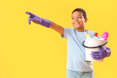 Küçük Afro-Amerikalı çocuk kovada temizlik malzemeleriyle sarı arka planda bir yeri işaret ediyor.