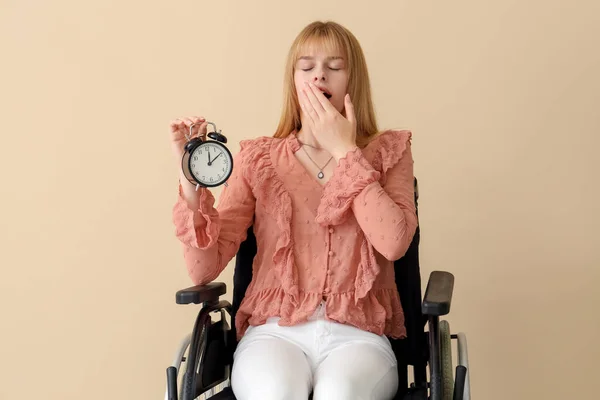 Sleepy young woman in wheelchair with alarm clock on beige background