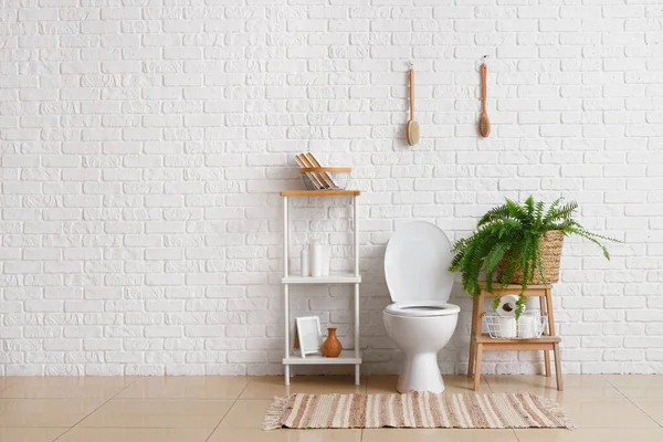 stock image Interior of light restroom with ceramic toilet bowl, shelving unit and houseplant near white brick wall