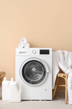 Çamaşır makinesi, bornozlu tabure ve bej duvarın yanında deterjan şişeleri.